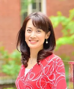 Smiling woman in a red patterned blouse posing outdoors with greenery and a brick wall behind her.